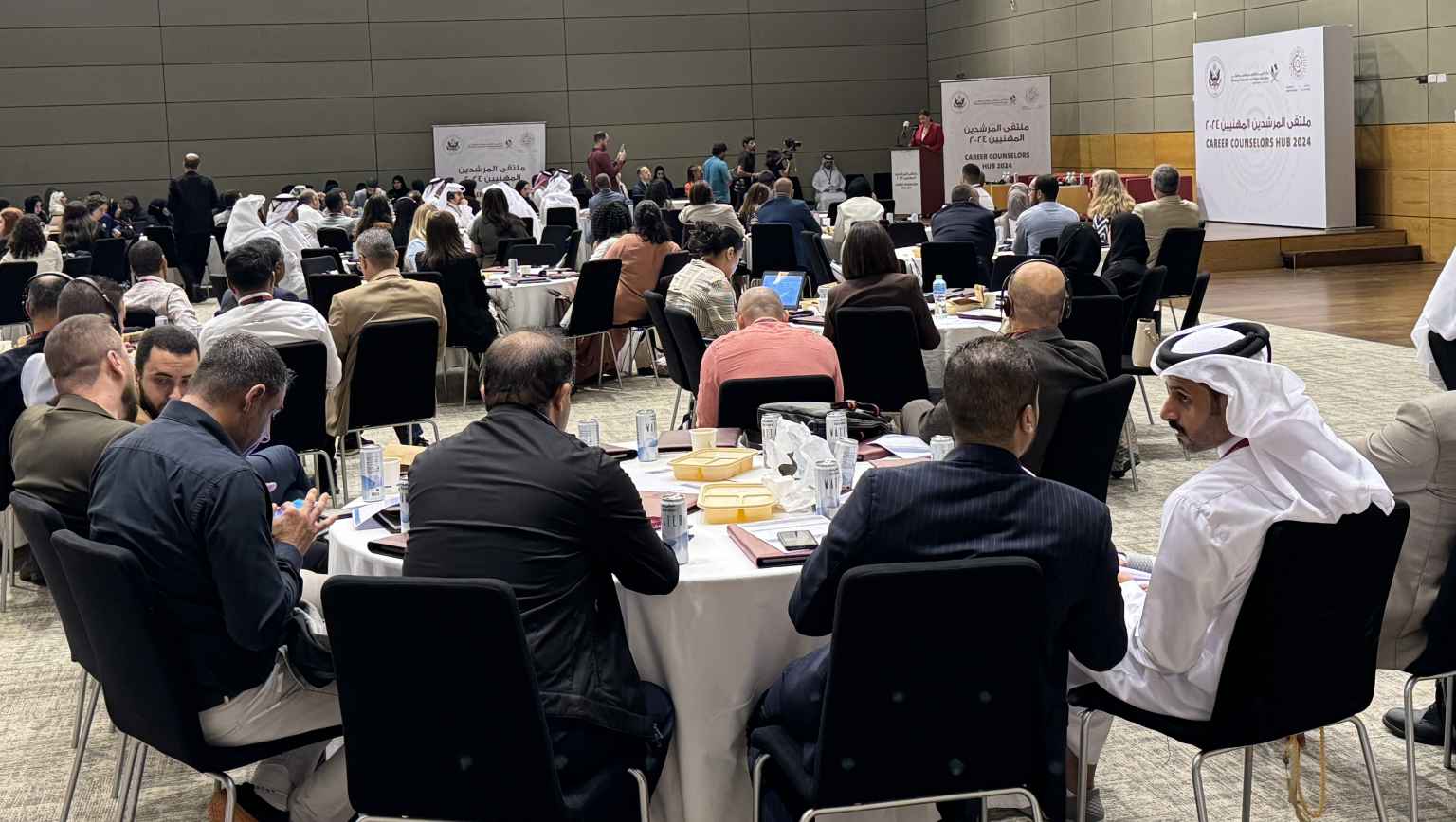 An audience of school counselors sitting in a ballroom listening to welcoming remarks by the Deputy Chief of Mission at the U.S. Embassy in Qatar